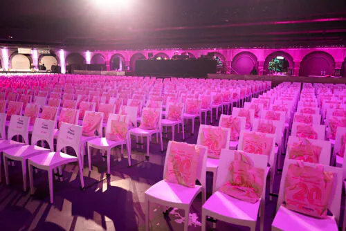 Salle vide avec des rangées de chaises blanches et des sacs cadeaux sous une lumière rose.