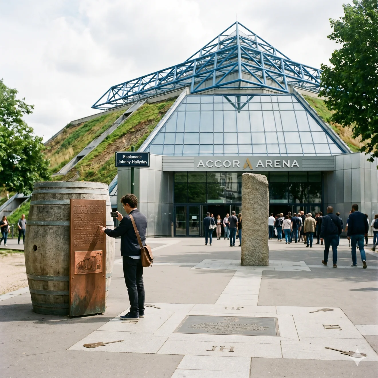 Un homme de dos regarde un panneau historique en forme de tonneau devant la structure pyramidale inclinée et végétalisée de l'Accor Arena.